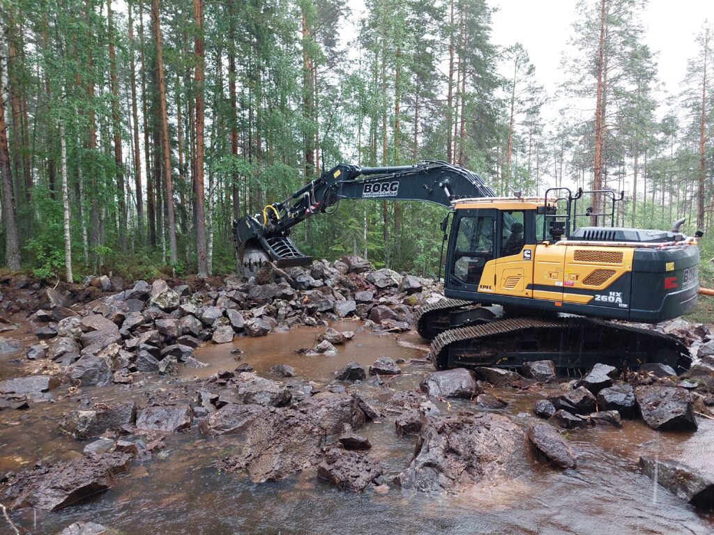 En grävmaskin gräver i sidan av ett vattendrag, där det ligger stora mängder sprängd sten.