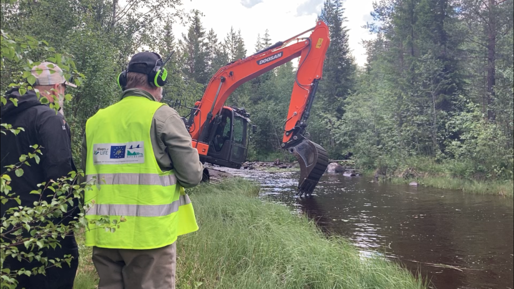 Två män med ryggen mot kameran står framför en grävmaskin i vatten.